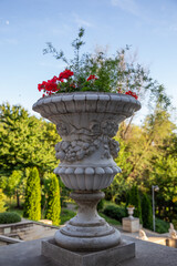 A decorative stone vase with red flowers and greenery standing on a terrace in Valea Morilor Park, Chisinau, Moldova. The ornate classical design, soft natural light, and lush green background create 