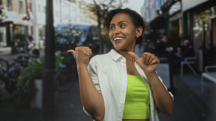 Woman in white shirt over neon green garment pointing thumbs to herself on a busy street cafe with bicycles and lamppost visible; confidence.