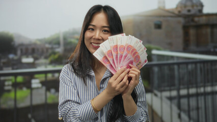 Young chinese woman holds yuan banknotes with a smile, set against the backdrop of ancient roman ruins, blending cultural symbols of finance and history in an outdoor setting.