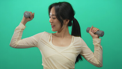 Young woman with dumbbells exercising against green wall, smiling confidently and showcasing fitness and strength.