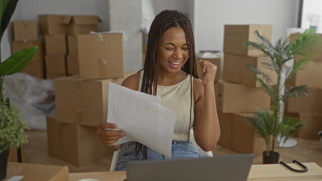 Woman holding papers and smiling while reading documents and making a fist pump in a building stacked with moving boxes; new home joy.