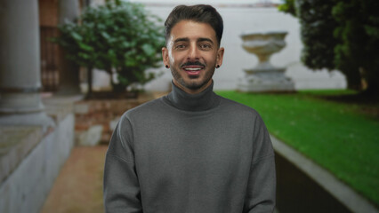 Fototapeta premium Young man face smiles toward camera near stone column in lush green park with blurred urn and foliage; joy.