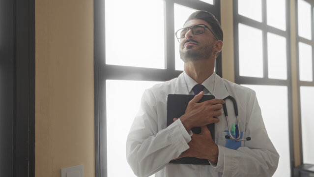 Man with stethoscope examines tablet while standing by window in clinic; dedication professional care.