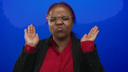 Naklejka premium African american woman covers eyes with hands while wearing glasses and red blouse in a blue studio; shyness.