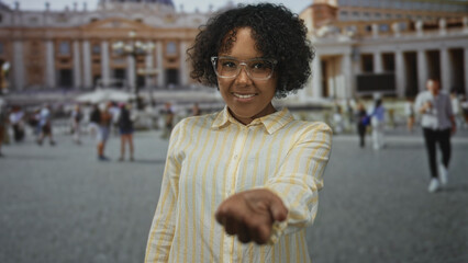 Woman with glasses smiles and holds out palm on street in st peter's square at vatican building;...