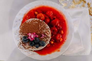 Delicious dessert featuring a round cake topped with cocoa powder, fresh blueberries, and pink flowers, served with vibrant strawberry sauce on a white plate with elegant presentation