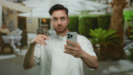 Young man with beard using smartphone on terrace of outdoor restaurant in city capturing candid moment of surprise and communication in urban setting.