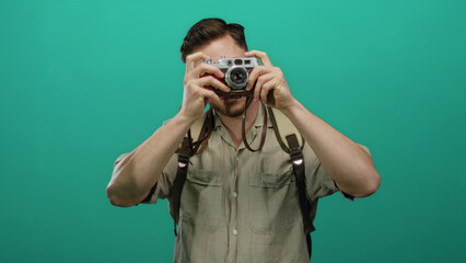 Young man with beard holding camera against a green wall, embodying a tourist photographer vibe with backpack, wearing casual attire, amid an isolated background.