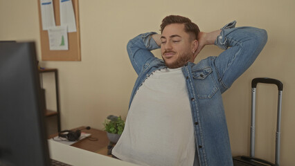 Young man with beard working at desk in office setting wearing denim shirt focused on computer screen showcasing a professional work environment indoors.