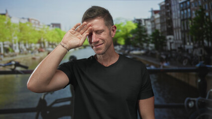 Man with hand to forehead shielding eyes on amsterdam street canal bridge, wearing black tshirt and...