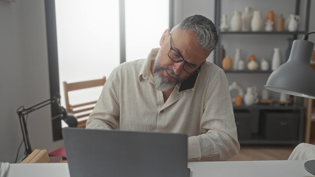 Man with glasses and gray beard holding phone to ear while typing on laptop at desk in a building office workspace; stress deadline.