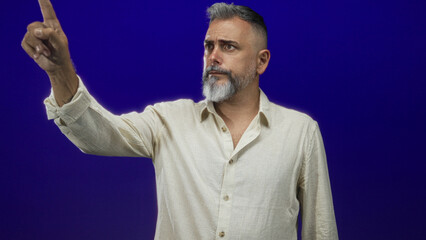Middle aged man points index finger with a serious expression in a deep blue studio wearing a beige linen shirt; authority.