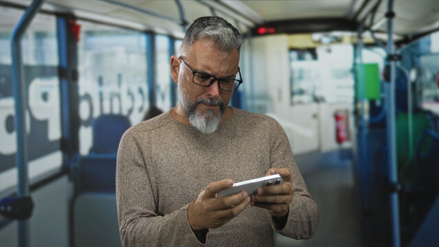 Man with hand on neck for neckache holding smartphone on bus, wearing sweater and glasses; commute frustration.
