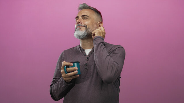 Middle aged man with gray beard holding teal mug and touching ear while smiling in pink studio; contentment reflection thought.