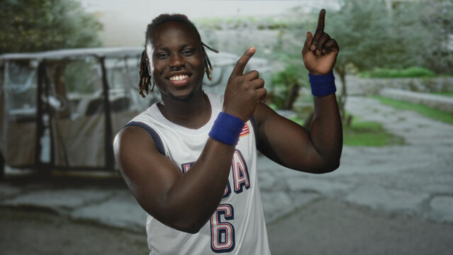 Man in usa basketball jersey number 6 pointing finger upward with both hands and smiling in street near golf cart; joyful confidence.