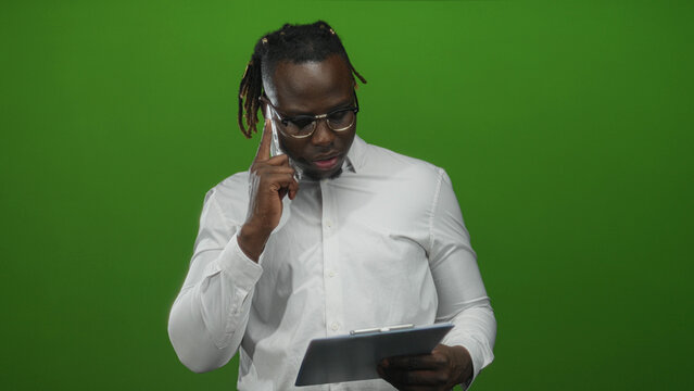Young african american man holds phone to ear while studying a tablet and clipboard in a green studio under soft lights; serious planning. - Powered by Adobe