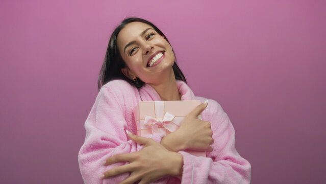Hispanic woman joyfully holding a pink gift box while wearing a robe against a vibrant pink background wall, radiating happiness and excitement.