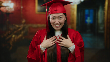 Young woman in red graduation gown celebrating with joy in an indoor museum setting, expressing happiness and accomplishment surrounded by elegant decor.