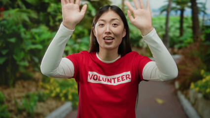 Obraz premium Woman wearing red volunteer shirt with arms raised smiling in a lush park setting showcasing joy and engagement in outdoor activities