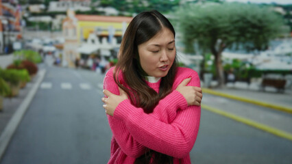 Fototapeta premium Woman in pink sweater in urban street setting with arms crossed appearing cold with blurred cityscape background.