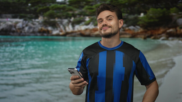 Man holding smartphone and looking at the screen on a sandy beach shore near turquoise water; calm contemplation.