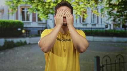 Man covering eyes with hands wearing yellow shirt that reads happiness in front of building; joyful playfulness.
