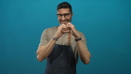 Man with hands forming heart in teal studio, wearing denim apron and glasses, smiling at camera; affection and connection.