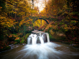 Beautiful waterfall cascades under a charming bridge in Luxembourg during autumn