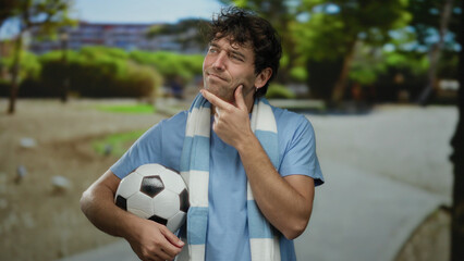Hispanic man in park holds soccer ball, contemplating thoughtfully, surrounded by green scenery and showcasing athletic appeal in a sporty, outdoor setting.