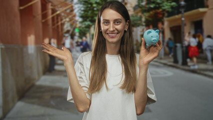 Woman smiling while holding a blue piggybank on a busy street, palm up and open hand showing...