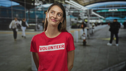 Woman wearing volunteer shirt smiling with eyes closed and hands behind back at airport terminal; helpfulness serenity community.