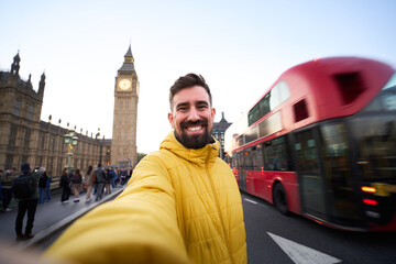Smiling caucasian man taking a selfie in London with Big Ben and a red double decker bus passing behind. Wide angle portrait on Westminster Bridge captures city energy, travel joy and urban vibes.
