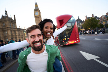 Joyful interracial couple having fun in London, with Big Ben and a red double decker bus behind. Playful piggyback pose, wide angle travel portrait that radiates energy and spontaneity.