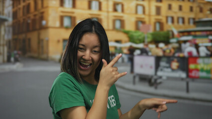Young chinese woman in green volunteer t-shirt smiling and gesturing energetically on a bustling city street with vibrant buildings and blurred passersby in the background.