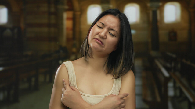 Young chinese woman with an expressive face stands indoors in a church setting, arms crossed, conveying confusion or displeasure, highlighting cultural intersection and emotional depth. - Powered by Adobe