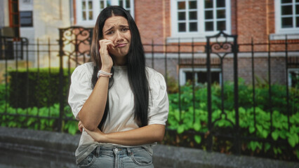 Fototapeta premium Young hispanic woman wiping tears with hand, arm crossed, standing by iron fence in front of building; sadness vulnerability isolation.