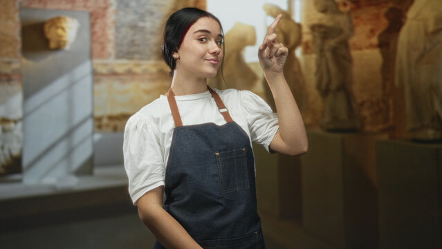 Woman in apron pointing finger upward in a museum building, hand gesture guiding a tour and looking up; curiosity guidance.