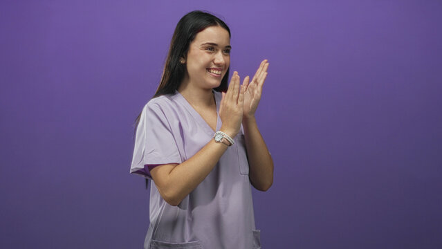 Young nurse woman clapping hands and smiling in a purple studio setting, wearing lavender scrubs and a wristwatch; joy gratitude.