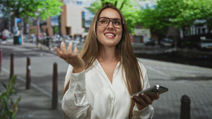 Woman smiling while texting on smartphone on city street, wearing glasses and white shirt; everyday happiness.