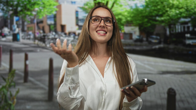 Woman smiling while texting on smartphone on city street, wearing glasses and white shirt; everyday happiness.
