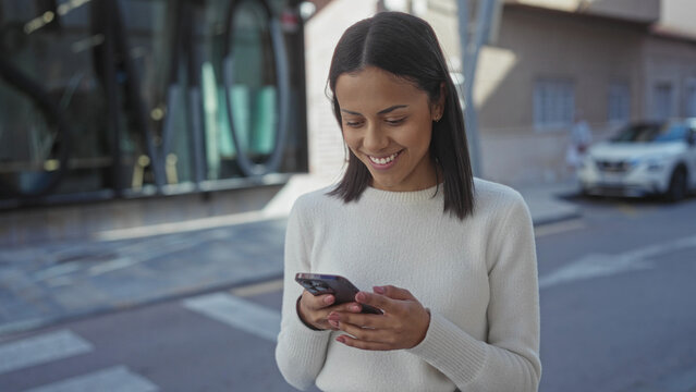 Young hispanic woman using smartphone on sunny street outdoors, smiling confidently as she engages with technology in an urban environment. - Powered by Adobe