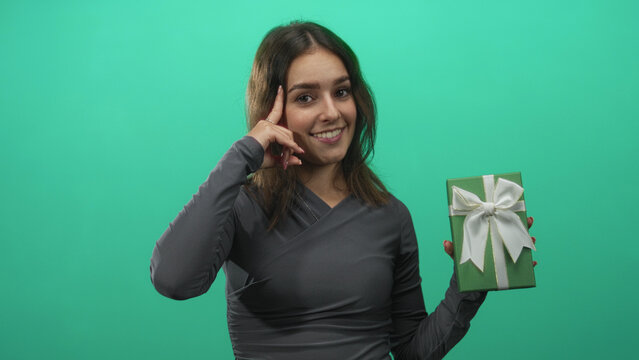 Woman holding green gift box with finger to temple gesture against green studio wall; thoughtful surprise idea.