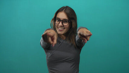 Woman wearing glasses points both index fingers toward camera in studio with teal background; playful invitation.