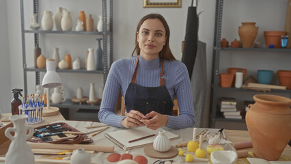 Woman shaping clay with hands at a ceramics studio table surrounded by pottery tools and vases on shelves; calm concentration.