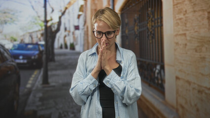 Woman with glasses touching chest and bringing palms together in a breathing exercise on a cobbled city street near iron gate and parked cars; mindfulness calm.