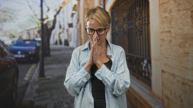 Woman with glasses touching chest and bringing palms together in a breathing exercise on a cobbled city street near iron gate and parked cars; mindfulness calm.
