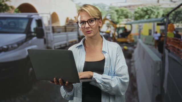 Woman holding laptop and typing with hands while wearing glasses and a light shirt amid trucks and excavator on street construction site; site management focus.