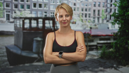 Woman with arms crossed showing smartwatch in sports bra and leggings in front of building by canal; confidence determination.