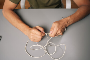 Close-up of technician hands tying knot in damaged white charger cable, attempting repair at gray...