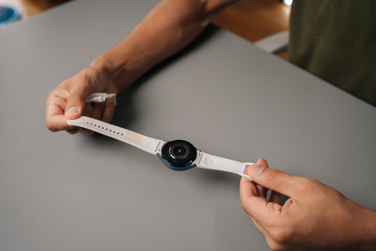 Top view of unrecognizable man maintaining personal device hygiene, disinfecting modern smartwatch with antibacterial spray, sitting at table. Concept of healthy lifestyle.